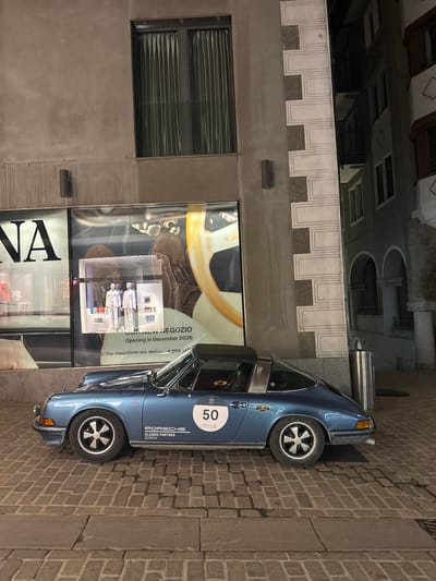 A classic light blue Porsche 911 Targa is parked at night on a cobblestone street in Sankt Moritz, Switzerland. The vehicle, facing left, features a black targa roof, Fuchs-style wheels, and a white circular "50" decal on the passenger door. "PORSCHE CLASSIC PARTNER ZURICH" is stenciled along the lower body, with a "CHOPARD" sticker above it. To the left, a modern building with a large storefront window displays two mannequins dressed in light clothing. Text visible on the window indicates "OUR NEW NEGOZIO Opening in December 2026" and partially "GIANNA". Above the storefront, the building has a plain, light-colored facade with a dark-framed window featuring vertical blinds. Water streaks are evident on the upper wall. To the right of the parked car, a building with traditional alpine architecture is visible, characterized by decorative white quoins against a darker facade and arched elements on lower levels. A stainless steel bollard or waste receptacle stands near the car's rear. No individuals are present in the scene.