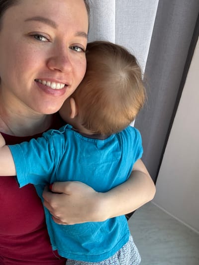 A close-up, portrait-oriented image depicts an adult female holding a young child indoors in Gamovo, Russia. The adult female, positioned on the left side of the frame, has light-colored eyes, light skin with visible freckles, and a discernible smile revealing her upper teeth. Her hair is pulled back, and she wears a dark red top. A faint reddish mark is present on her neck below her left ear. Her left hand, with red-painted fingernails, supports the child. The child, an infant or toddler, is seen from behind, with reddish-blonde hair and the back of their head resting against the adult's face or shoulder area. The child is dressed in a short-sleeved, bright blue t-shirt with a ruffled collar and light-colored, patterned shorts. The background features vertical panels of light-grey and dark-grey curtains or blinds, with a plain white wall section visible on the far right. Natural light illuminates the subjects from the right.