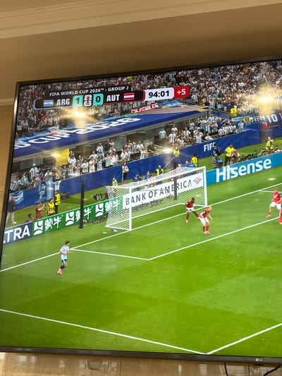 This image depicts a television screen displaying a soccer match. The score shows Argentina (ARG) leading Austria (AUT) 1-0. The match is in the 94th minute with 5 minutes of added time. The game is identified as FIFA World Cup 2026™, Group J. The screen shows the green turf of a soccer field, with players in red and white jerseys in action near a goal. Spectators are visible in the background, wearing white and light blue attire, suggesting support for Argentina. Commercial advertisements for "BANK OF AMERICA" and "Hisense" are displayed on the screen. The television is set on a tiled surface, presumably in a residential setting in North Bergen, United States of America, given the context of the provided location.