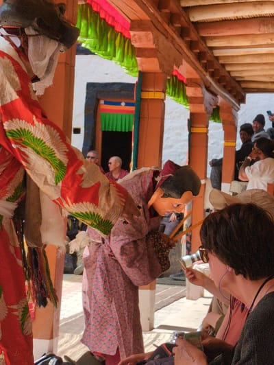 In Pibiting, India, a traditional masked dance performance takes place under a wooden canopy adorned with colorful fabric streamers. Two individuals in elaborate costumes are the central figures. One, on the left, wears a large, dark, animal-like mask and a vibrant red robe decorated with white and green feather-like patterns. The other, on the right, is partially visible, wearing a patterned pink robe and a tan mask with dark hair, holding two thin wooden sticks. They appear to be engaged in a ritualistic dance, with the masked figure on the right seemingly in motion. A crowd of onlookers is gathered behind them, observing the performance. Among the spectators, a person in the foreground is wearing sunglasses and holding a small object. The environment suggests an outdoor courtyard or temple setting, with white walls and wooden pillars supporting the roof structure. The ground is paved with a textured surface.