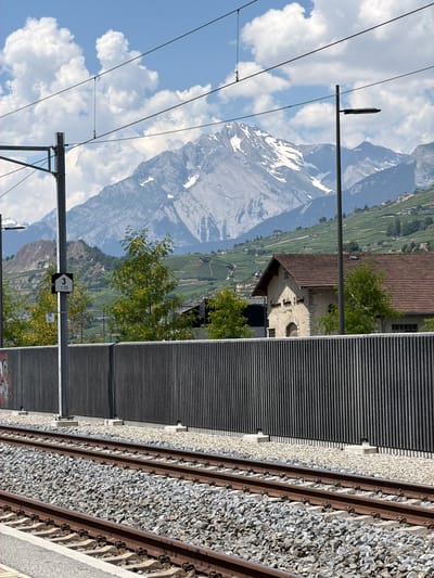 The image depicts a view from a train station platform in Saint-Léonard, Switzerland. In the foreground, two sets of train tracks are visible, laid on a bed of gravel. A gray metal fence runs along the side of the tracks, with a signpost showing "3 170" to the left. Beyond the fence, a building with a red tiled roof and stone walls is partially visible, with trees in front of it. In the background, rolling hills covered in vineyards ascend towards snow-capped mountains under a bright blue sky with scattered clouds. The scene is captured on a sunny day. There are no people visible in the image.