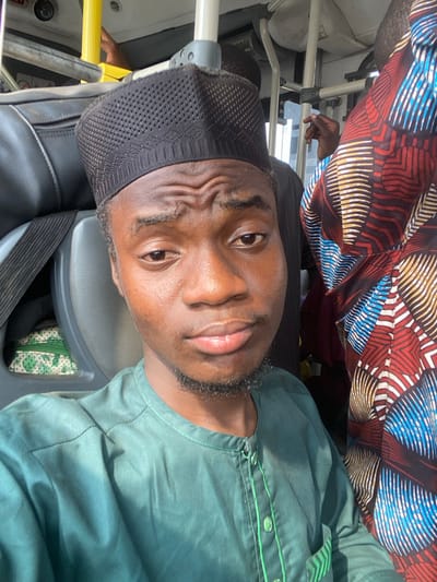 The scene depicts the interior of a passenger vehicle, likely a bus, in Ebute-Metta, Nigeria. A young Black male, approximately 18-25 years old, occupies the central foreground, looking directly at the viewer with furrowed eyebrows and a neutral to slightly concerned expression. He wears a dark, perforated skullcap (kufi) and a long-sleeved dark green tunic with a banded collar and green button placket. To his right, partially visible, is another individual wearing a vibrant, multi-colored patterned garment with red, blue, white, and black geometric designs. A dark-skinned hand is seen gripping a grey overhead support bar near this individual. The vehicle's interior features grey seating components, a yellow overhead grab handle, and grey metallic structural poles and rails. A light green and white patterned fabric object is partially visible behind the central subject's left shoulder.