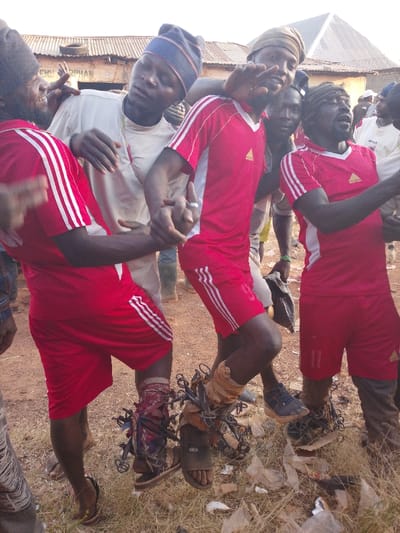 A group of men are closely gathered, some wearing red and white Adidas soccer jerseys and shorts. Their footwear varies from sandals to sneakers, some tied together with wire. One man in a white shirt and striped hat is being held, while another is making a hand gesture. The ground is littered with plastic bags and debris. A building with a sign is in the background.