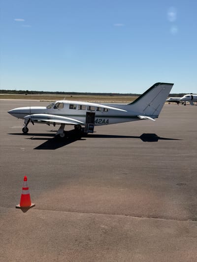 The image depicts a white and green twin-engine propeller aircraft parked on an asphalt tarmac under a clear blue sky. The aircraft's registration number, "42AA", is visible on the tail. The passenger door is open, with steps extended. A red and white traffic cone is positioned in the foreground to the left of the aircraft. In the background, other aircraft are visible, and a line of trees and vegetation forms the horizon. The scene appears to be an airport, likely in a suburban or rural area.