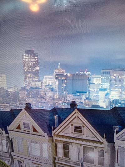 The image displays a nighttime cityscape, dominated by illuminated skyscrapers. In the foreground, rows of Victorian-style houses with distinct architectural details, including triangular gables and ornate window frames, are visible. An object with a bright core is positioned high in the sky. The overall mood is evocative, capturing the contrast between historic residential structures and the modern urban skyline.
