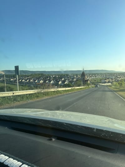 The image shows a view from inside a vehicle traveling on a paved road. The landscape outside the vehicle includes a settlement of houses with pitched roofs, a church with a tall spire, and a backdrop of distant hills. The sky is clear and blue. Another vehicle is visible on the road ahead. The interior of the vehicle includes parts of the dashboard, windshield, and possibly a side pillar. The location is likely Vlast Truda, Russia, based on the prompt.