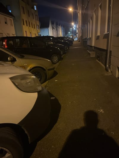 The image displays a nighttime urban street in Tuttlingen, Germany. A line of parked vehicles, primarily dark-colored, occupies the left side of the thoroughfare, receding into the background. In the foreground on the left, the front quarter of a white vehicle with a black bumper is visible, followed by a gold-colored sedan, and then multiple black compact cars. Multi-story residential-style buildings flank both sides of the street. The building on the left is light-colored with several windows, one showing internal illumination on an upper floor. The building on the right presents a dark stone-clad base, lighter upper stories, and a vertical rain gutter. A tall streetlight in the mid-ground provides the primary illumination, casting a distinct human shadow onto the foreground pavement. The paved surface is worn and displays scattered minor debris.