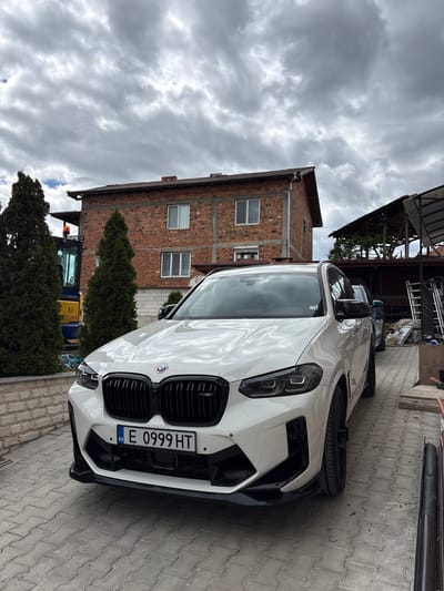 A white BMW X4 M Competition is parked on a brick paved driveway. Behind the car is a two-story brick building with three windows. Two tall evergreen trees stand in front of the building, partially obscuring a blue and yellow mini excavator. The sky is overcast with heavy, gray clouds. The license plate of the BMW reads "E 0999 HT" and has a Bulgarian flag on the left side. The scene is located in Dolno Drianovo, Bulgaria.