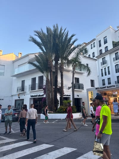 The image shows a street scene in Nueva Andalucía, Spain, during what appears to be late afternoon or early evening due to the warm, low sunlight casting soft shadows. Several people are walking across a pedestrian crossing. The buildings are whitewashed, characteristic of Mediterranean architecture, with balconies and awnings. Palm trees are interspersed among the buildings, adding to the tropical atmosphere.
One person in the foreground is wearing a bright neon yellow t-shirt and grey shorts, carrying a gold shopping bag. Next to them, another person wears a pink t-shirt and black shorts. Further down the street, a group of people are also crossing the street or walking along the sidewalk. Some are dressed casually in t-shirts, shorts, and pants. The overall mood seems relaxed and leisurely, suggesting a typical evening stroll or shopping excursion.
Visible text includes "MC2 SAINT BARTH" on a store facade and "SabadellSolbank" on another building, indicating the presence of retail establishments and a bank. The clear blue sky suggests pleasant weather. The scene captures a snapshot of everyday life in a popular tourist or residential area, with a blend of modern retail and traditional architecture.
