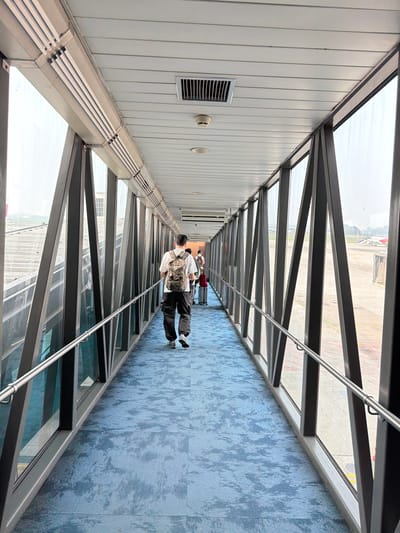 The image depicts a person walking down an airport jet bridge, a covered walkway connecting a terminal to an airplane. The jet bridge has large glass windows on both sides, offering views of the tarmac and distant buildings. The floor is covered with a patterned blue carpet. The person in the foreground is wearing a camouflage backpack, a white t-shirt, and black pants. Other individuals and luggage can be seen further down the jet bridge. The environment suggests a typical airport setting, likely in Rawa Burung, Indonesia, given the context.