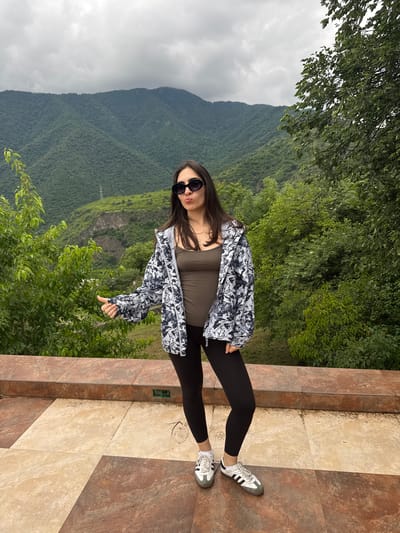 A young woman with long dark hair and sunglasses stands on a tiled patio with a mountainous, tree-covered landscape in the background. She is wearing a dark tank top, a patterned jacket, black leggings, and white sneakers. Her left arm is extended with her thumb up in a hitchhiking gesture. The woman has her lips puckered. The location is Akhtala, Armenia.