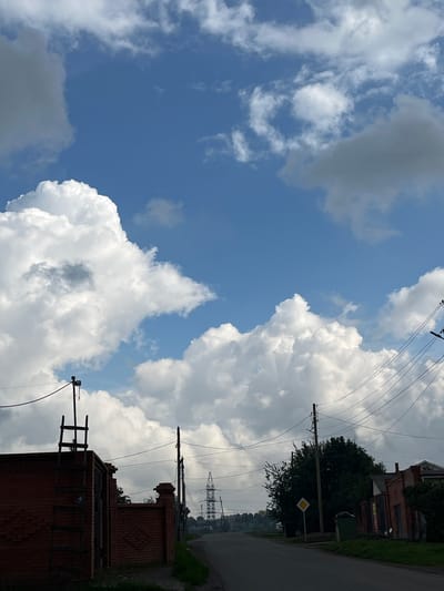 The image captures a street scene in the city of Сухая Балка, Russia, under a partially cloudy sky. In the foreground, a low, red brick wall extends across the left side of the frame. Leaning against the wall is a tall, weathered wooden ladder, reaching upwards towards a utility pole with electrical wires. The road curves gently into the distance, bordered by grassy verges and more brick structures on the right. Power lines crisscross the sky, supported by multiple poles. Further down the road, a tall lattice tower is visible on the horizon, suggesting a more industrial area. The sky is dominated by dramatic, white cumulus clouds against a bright blue backdrop, with some darker, more diffused clouds also present. A yellow diamond-shaped road sign is mounted on a pole near the right side of the road.