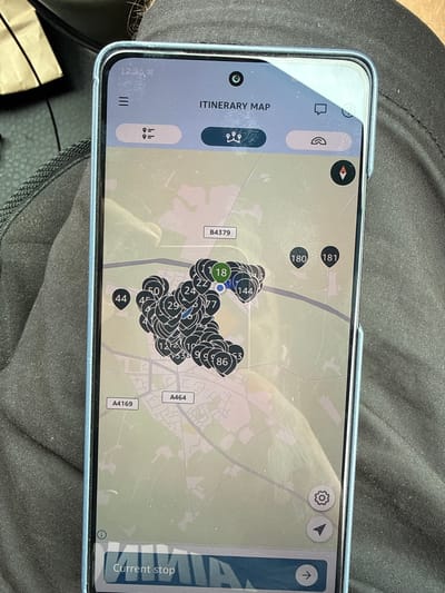 The image shows a smartphone displaying an itinerary map, likely of the city of Shifnal, United Kingdom, given the road labels A4169 and A464. The map is overlaid with numerous numbered circular markers, indicating various stops or points of interest. Some of the visible numbers include 1, 18, 22, 24, 29, 44, 50, 77, 86, 144, 180, and 181. The phone is positioned in a person's lap, with the screen facing upwards. The background consists of dark fabric, possibly clothing or upholstery, and a partially visible brown paper bag on the left side of the frame. The phone's interface includes icons for a menu, a chat bubble, a rainbow, and a compass. The bottom of the screen displays "Current stop" with an arrow, suggesting navigation functionality. The time displayed on the phone is 12:36.