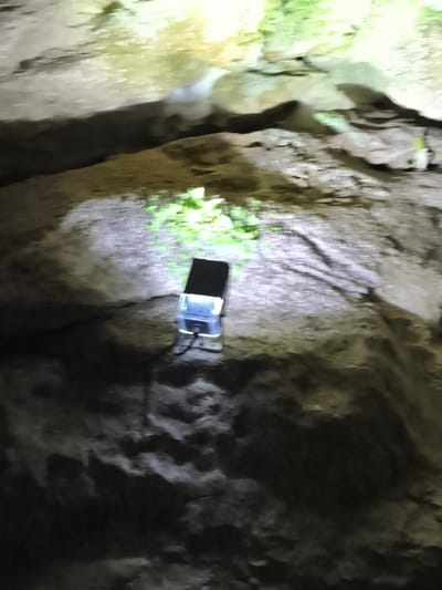 A black object with a clear plastic cover and a black top is placed on a textured, rocky surface. The object is illuminated by a bright light, casting shadows. In the background, more textured rocks are visible, with some areas lit by a green light source. The overall scene appears to be within a cave or an outdoor rocky environment. This image was captured at the Balinger Felsen, a natural rock formation located in Hayingen, Germany.
