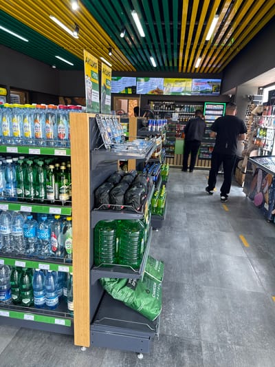 The photo shows a grocery store aisle with shelves stocked with various items, including bottled water, charcoal, and other goods. Two men are walking down the aisle. The walls are decorated with yellow and green stripes. The store appears to be modern with gray tiled floors.
Several signs marked "АКЦИЯ" in cyrillic appear on the shelves, suggesting a sale. The presence of brands with cyrillic labels indicates the location is likely in a country where Russian is spoken or understood.
The inclusion of brands such as "CORONA" and the fact that Kyrgyzstan labels include products in English suggest this store is in Кашат, Kyrgyzstan.