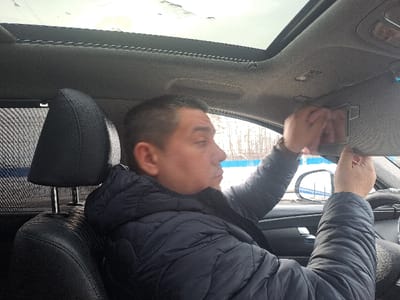 An adult male is seated in the driver's seat of a vehicle, positioned in Obukhovo, Russia. He is observed in profile, facing right, and interacting with the car's sun visor mirror. He wears a dark blue puffer jacket. His right hand supports the sun visor, while his left hand is positioned over the mirror's surface, appearing to wipe or adjust an object within it. The vehicle interior features a black driver's seat and headrest, a light-colored headliner, and a visible sunroof. A sunshade is partially visible on the rear side window. Through the car windows, an outdoor environment is discernible, characterized by bare trees, a blue and white striped barrier, and ground covered in snow or ice, under an overcast sky.