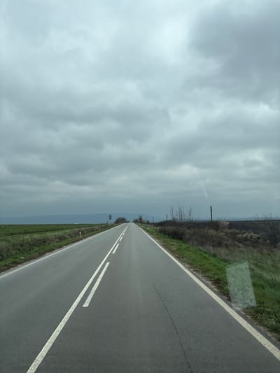 The image depicts a road in Pamukchii, Bulgaria, under a cloudy sky. The asphalt road has two white dashed lines in the center and a solid white line on the right edge. The roadside vegetation is sparse, consisting of grass and shrubs. There are power lines present along the road.