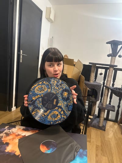 A woman with dark hair and bangs holds a blue and yellow marbled vinyl record that reads "Tristesse Business" in front of her. Behind her, partially obscured by a wire rack, is a cat tree. The room features a black door and a white wall. To the side, vinyl record sleeves with abstract artwork are visible.