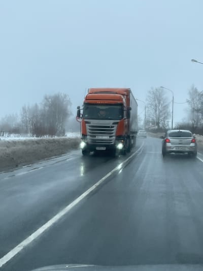 The image depicts a two-lane asphalt road in Pokrovskoe, Tver Oblast, Russia, under overcast and hazy winter conditions. The road surface is wet, indicating recent precipitation or melting snow. An orange Scania semi-truck, featuring a white box trailer and illuminated headlights, occupies the oncoming lane, moving towards the viewer. Its Russian license plate displays X538A A 69. In the same lane as the perspective of the camera, a silver Chevrolet Cruze hatchback travels away from the viewer, showing Russian license plate M 361 MH 69. A third, less distinct, light-colored vehicle is visible further ahead in the distance. The left roadside exhibits a snow-covered verge and a line of bare deciduous trees obscured by fog. The right roadside features additional bare trees and utility poles. The sky is uniformly light grey and appears foggy. No pedestrians or individuals are visible outside the vehicles.