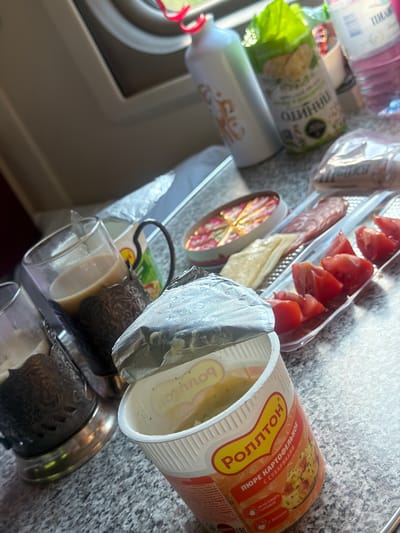 An overhead shot captures a tray of food and drinks, likely from a train or roadside establishment. A cup of instant mashed potatoes, labeled "РОЛЛТОН" and "ПЮРЕ КАРТОФЕЛЬНОЕ С СУХАРИКАМИ," is open in the foreground, with its foil lid partially peeled back. To the left, two metal cup holders contain glasses of a light-colored beverage, possibly tea or coffee. The tray also holds sliced tomatoes, a piece of cheese, and what appears to be salami. In the background, a bottle of water, a white bottle with an orange design, and a green package of snacks are visible. The overall setting suggests a travel meal, possibly in a compartment with a window showing a blurred outdoor scene. No people are present in the image.