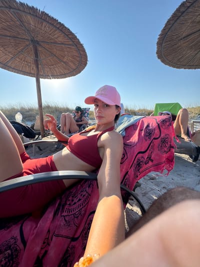 A woman with a pink baseball cap and a burgundy athletic top sits in a beach chair on a sandy beach, with dry grass in the background under a clear blue sky. She is looking towards the camera and has a hand resting on her thigh. A pink towel with a black pattern is draped over her chair. To her right, another person is visible in the background, also sitting in a beach chair with a green covering. Two thatched umbrellas provide shade. The scene appears to be in the Municipal Unit of Moudros, Greece.