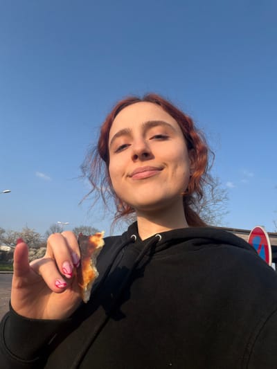 A young woman with reddish-brown, wavy hair, wearing a black hooded sweatshirt, is depicted in an outdoor setting in the city of Toul, France. She is seen from the chest up, looking directly at the camera with a faint smile. Her left hand, adorned with pink fingernails featuring white swirl patterns, holds a partially eaten piece of food, resembling a bread crust or pizza edge. The background features a clear blue sky with minimal clouds. To the left, two grey streetlights are visible, along with bare trees and some white blossoms. To the right, a circular red and blue road sign, consistent with a "no stopping" or "no parking" symbol, is partially observed behind the woman. The ground visible in the lower left background appears to be a paved surface.