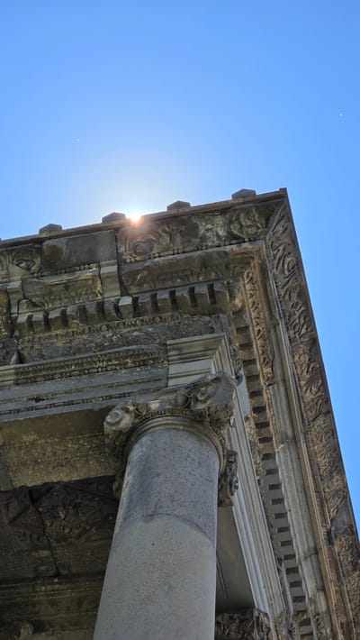 The image captures a low-angle view of the Garni Temple in Garni, Armenia, against a clear blue sky. The sun is visible, peeking over the edge of the ancient stone structure, casting a bright glare and lens flare. The temple exhibits classical architectural elements, including a fluted column and elaborately carved stone friezes and cornices. The stonework appears weathered and aged, with visible cracks and signs of wear. The details of the carvings, featuring intricate patterns and motifs, are discernible despite the shadows and the bright sunlight. The overall impression is one of historical grandeur and enduring architecture.