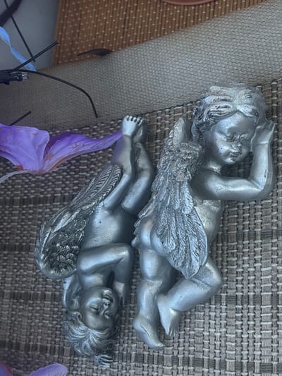 This image shows two silver cherub figurines lying on a textured, woven surface. One cherub is lying on its back with its head resting on its arm, and the other is lying on its stomach with its legs in the air. A purple artificial flower petal is visible in the foreground. The background appears to be a wicker mat and a brown surface with a dotted pattern. There are no people or animals visible in the image. The specific location in Osterode am Harz, Germany cannot be determined from the provided image.