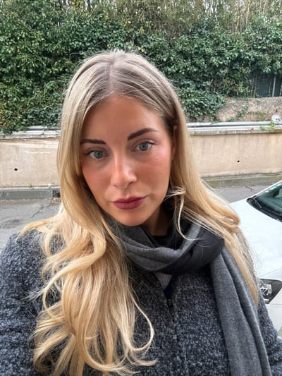 A blonde woman with blue eyes and dark red lipstick is depicted in a close-up outdoor portrait in Aix-en-Provence, France. She has long, wavy blonde hair and is wearing a dark grey textured coat with a dark grey scarf. Behind her, a light brown stucco wall is visible, topped by a dense wall of green ivy. Above the ivy, thin, bare branches or trees are present. To the woman's right, the front section of a white vehicle is partially visible. A dark asphalt surface is also discernable on the lower left.
