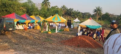 The image depicts an outdoor event taking place in Alayi, Nigeria, set on dry, open ground bordered by dense green vegetation and palm trees. On the left, a dark green SUV is parked next to a series of three interconnected rectangular tents, colored in red, blue, yellow, and teal sections. Under these tents, several individuals are seated on white plastic chairs. In the center, a larger, round tent with yellow and teal stripes features a decorated archway composed of white pillars and greenery; a man in a white suit stands beneath it near a yellow-draped table. Two large black audio speakers are positioned behind this central tent. To the right, additional white and yellow-accented tents shelter a larger group of seated attendees, including adults and children. A prominent mound of reddish-brown soil is situated in the mid-ground to the right. In the immediate foreground, partially visible on the right, is a man wearing a patterned blazer and a black baseball cap.
