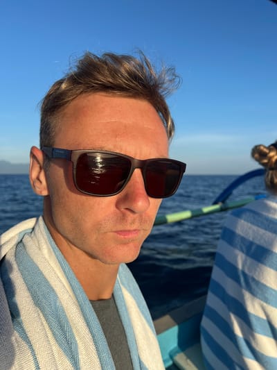 A medium shot shows a man wearing sunglasses and a blue and white striped towel on a boat. The background includes a body of water and a blue sky. The location is Seririt, Indonesia.