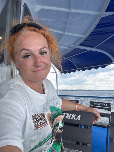 A woman with reddish-blonde hair pulled back and sunglasses perched on her head is seated on a boat. She is wearing a white t-shirt with graphics on it and has blue eyes and fair skin with freckles. She is smiling slightly at the camera. The boat has a blue awning overhead. In the background, a body of water is visible with a cityscape on the horizon. The name "БАЛТИКА" is visible on the back of the seat in front of her, which is a brand of Russian beer. The location is identified as Grebeni, Russia.