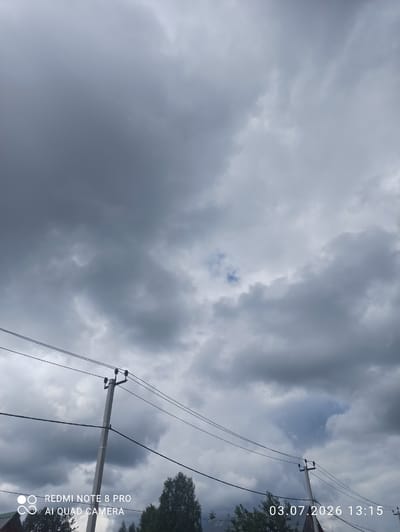 An outdoor, medium shot photograph depicts a cloudy sky with power lines and utility poles in the foreground. The sky is filled with dark, heavy clouds, suggesting overcast or stormy weather. Two utility poles are visible, with several power lines strung between them. The poles are grey and appear to be made of concrete. Below the power lines, the tops of trees and the roof of a building can be seen. A timestamp in the lower right corner indicates the date and time as "03.07.2026 13:15". In the lower left corner, a watermark displays "REDMI NOTE 8 PRO AI QUAD CAMERA". The image was taken in the city of Sovetskiy, Russia. There are no people or animals visible in the frame.