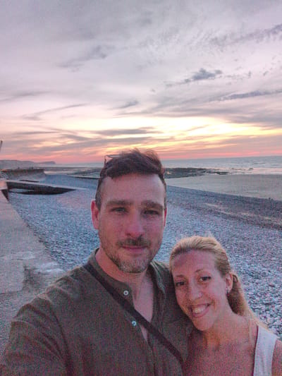 A selfie taken at dusk on a pebble beach shows a man and a woman standing close together. The man, with short dark hair and stubble, is wearing a dark green collared shirt. He is positioned on the left side of the frame. The woman, with blonde hair pulled back, is on the right, wearing a white top. She is smiling. Behind them, the sea stretches to the horizon, with a soft, colorful sky above, indicating sunset. The beach is covered in smooth, gray pebbles, and a concrete walkway runs along the left edge of the frame. In the distance, the coastline is visible with hills. The location appears to be Veules-les-Roses, France, given the characteristic pebble beach and the coastal landscape.