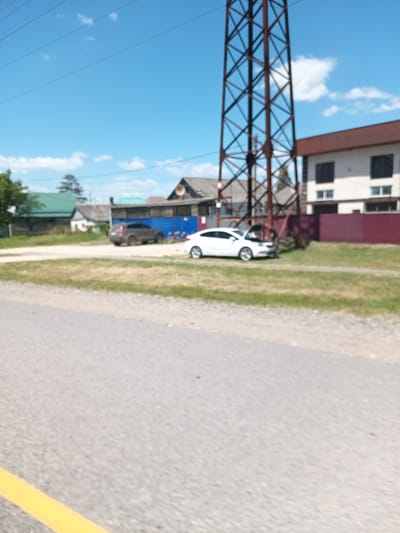 The image shows a residential area with houses, a power line tower, and two cars on a sunny day. A brown sedan is parked on the left side of the frame, and a white sedan with its hood open is parked on the right, with a person visible near the open hood. The ground is a mix of gravel and grass. The sky is blue with scattered clouds. This scene is located in the city of Krymsk, Russia.