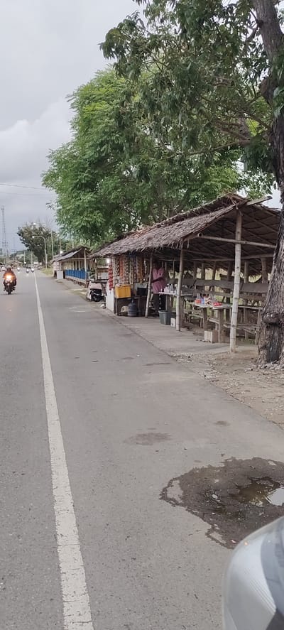 The image depicts a roadside scene in Keudealuerheung, Indonesia, characterized by a paved road with a white lane divider. To the right of the road are several small, open-air stalls with thatched roofs and wooden structures, likely for vendors. One stall is occupied by a person wearing a pink shirt, standing behind a display of goods, possibly snacks or small items. Tables and seating are present at some of the stalls. A motorcycle is visible on the road, moving away from the viewer. In the background, trees and vegetation line the roadside, and a telecommunications tower is visible in the distance. The sky is overcast, suggesting a cloudy day. The pavement shows some signs of wear, including a puddle on the right side.
