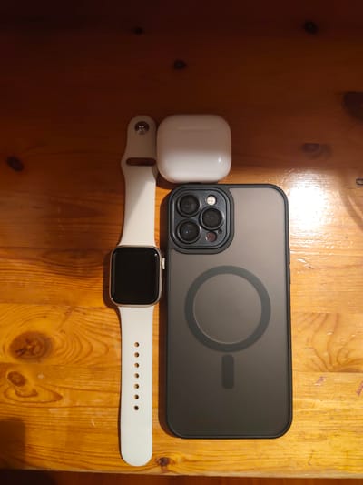 An Apple Watch, AirPods charging case, and a dark-colored smartphone case are arranged on a wooden surface. The Apple Watch has a white band and a black screen. The AirPods case is white. The smartphone case is dark with a circular design and a vertical line in the center, indicating it is compatible with MagSafe. The wooden surface appears to be a table with visible grain and some knots. The lighting suggests an indoor environment. The image does not contain any elements that would allow for identification of its location in Neckertal, Switzerland.