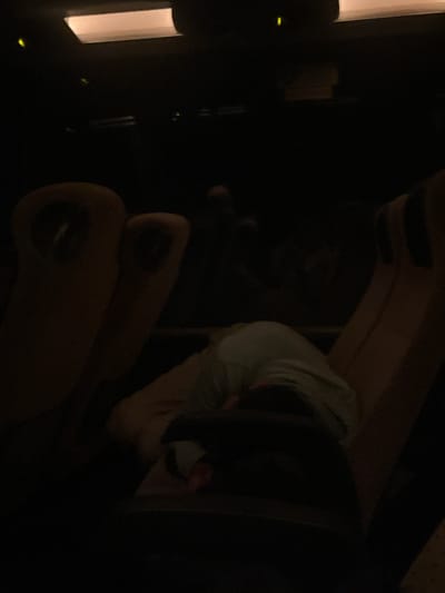 The image depicts a dimly lit interior scene within a cinema or theater located in the city of Iastrebino, Bulgaria. Multiple rows of upholstered seats, light brown or tan in color, are visible. In the foreground, center-left, an individual is seated, slumped forward with their head resting on their chest or lap. This person is wearing a light-colored top, possibly white or light blue/green, and dark lower garments. A small, indistinct red object is partially visible near their right side. To the immediate left and right of the foreground subject, adjacent empty seats are observed. In the mid-ground and background, further dark shapes suggest additional seating rows and occupants, with the faint outline of a head visible in the far background. Overhead, a rectangular, ceiling-mounted light fixture emits a soft, diffused glow. Several small, bright yellow-green indicator lights are scattered across the dark background, suggesting emergency exits or operational indicators. The overall environment is characterized by very low ambient light.
