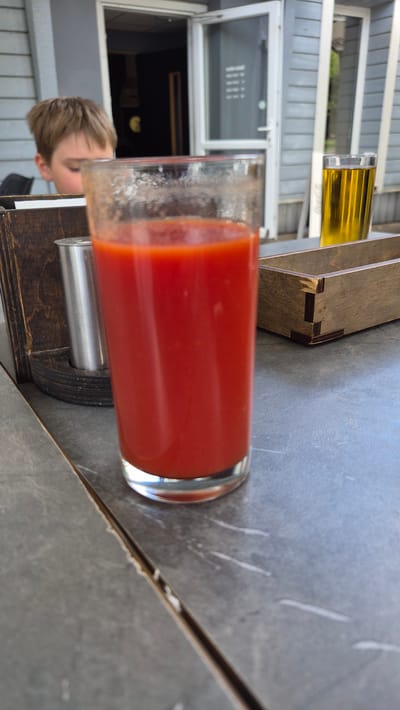 The image shows a close-up of a tall glass filled with a vibrant red liquid, likely tomato juice, sitting on a textured gray surface. To the left of the glass, a small portion of a person's head, specifically a child's blonde hair and part of their face, is visible, suggesting they are seated or standing near the drink. In the background, out of focus, a window with horizontal blinds and a building with gray siding can be seen, hinting at an outdoor or semi-outdoor setting. The lighting appears natural, suggesting it could be daytime. The overall composition focuses on the drink, with the person and background elements serving as secondary context. The scene appears casual and relaxed.