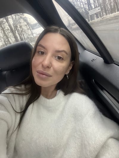 The image displays a female individual situated within a vehicle in Vysokovo, Russia. She is directly facing the camera, characterized by dark brown hair, light brown eyes, and full lips. She wears a white, knitted crew-neck sweater and a silver hoop earring with a cross pendant in her right ear. The vehicle's interior shows a black car seat headrest behind her left shoulder and a dark vehicle door panel to her right. Through the window, an exterior scene depicts a leafless, wintry or early spring landscape with bare trees and a wet, slushy road. A white rectangular sign, bearing black Cyrillic text, is visible and translates to "RESTAURANT 'AUGUST' -> 1.8 KM".