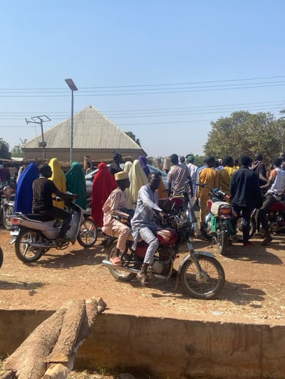 This image depicts an outdoor gathering in Konkiyel, Nigeria, under a clear, bright blue sky with strong sunlight casting distinct shadows.
In the immediate foreground, a low, weathered concrete barrier extends horizontally across the frame. Two large, cut tree logs with bark are positioned on the ground in front of this barrier, to the left.
The mid-ground is dominated by multiple individuals and motorcycles. From left to right: A person in a dark long-sleeved shirt and grey trousers sits astride a silver scooter-style motorcycle, facing right. Directly behind this motorcycle, a group of women are visible, wearing full-body coverings in bright colors including blue, yellow, green, and red, along with two in cream/beige. To their right, a red standard motorcycle is positioned, with a male driver in a light blue/grey traditional outfit and a male passenger (pillion rider) in a light brown traditional outfit and a matching cap, both facing right. Another standard motorcycle is partially visible to the right of this pair, facing slightly left.
The background features a dense crowd of individuals, predominantly male, standing and gathered. Visible among them are woven sacks, suggesting a market environment. To the left, a prominent building with light-colored, corrugated metal forming a distinct pyramidal roof is visible. A tall, grey pole with a solar-powered streetlight extends upwards to the left of this building. Overhead, three parallel power lines traverse the upper mid-ground. To the far right, a large tree with green foliage is visible. The ground throughout the scene is a reddish-brown dirt.