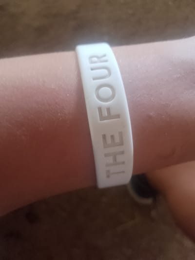 A white silicone wristband with the words "THE FOUR" embossed in raised capital letters is worn on a person's arm. The wristband is positioned horizontally across the arm, with the text oriented to be read from left to right. The arm appears to be lightly tanned and is in focus, while the background is blurred. The background consists of a reddish-brown textured surface, likely a floor or furniture, and hints of dark fabric, possibly clothing. The lighting suggests an indoor setting, and the overall image has a casual, everyday feel. No specific activity or event is depicted beyond the wearing of the wristband. The time of day and weather are not discernible. The location context of Inhulo-Kamianka, Ukraine is provided but not visually evident in the image itself.