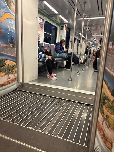 The image depicts the interior of a modern train car, likely part of the FGC (Ferrocarrils de la Generalitat de Catalunya) network, stopped at a station in Sant Quirze del Vallès, Spain. The view is from a slightly elevated perspective, looking into the main passenger compartment through an open inter-car doorway, which features a retractable metal plate with parallel slats covering the gap in the floor.
Two female passengers are prominently seated on grey, cushioned seats on the left side of the aisle. The woman in the foreground, with grey hair, wears a multi-colored striped jacket (blue, white, black), dark patterned pants, and pink and white sneakers. She is holding and looking at a smartphone. Next to her is a black bag. The woman further down the aisle has dark hair, wears a dark blue puffer jacket, dark pants, and light-colored sneakers, with her feet resting on the opposite seat. She is holding and looking at a tablet or large smartphone. A teal bag rests on the seat next to her.
Further down the train car, another passenger is partially visible on the right, seated and accompanied by a dark-colored cello in a case.
The train interior features white walls, overhead fluorescent lighting, and numerous silver vertical support poles and horizontal handrails. A red "SOS" emergency sign and other informational stickers are visible on the wall above the windows. Advertisements are displayed on the interior panels. The poster closest to the camera on the left depicts a paraglider with an orange parachute over a mountainous landscape with a lake, and a smaller image of a person cycling. Partially visible Catalan text reads "amb tu... gaudeix la tenim Tdt." A QR code is also visible on this poster. The floor inside the passenger compartment is light grey.
