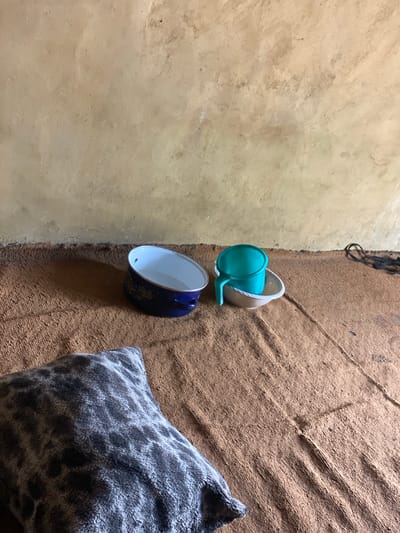 A low-angle, indoor shot shows a scene in what appears to be a room in Daniel, Nigeria. The background is a textured, light brown wall with some darker patches and an uneven surface. In the mid-ground, a brown, coarse-textured blanket covers the floor. On the blanket, to the left, is a blue enamel bowl with a floral pattern on its exterior. The bowl is partially filled with water. To the right of the blue bowl is a white saucer with a turquoise plastic mug resting inside it. The mug also contains water. In the foreground, the corner of a grey and black speckled throw pillow is visible, lying on the blanket. A black cable is visible in the background, partially obscured by the wall and blanket. The lighting suggests an indoor setting with natural light coming from an unseen source.