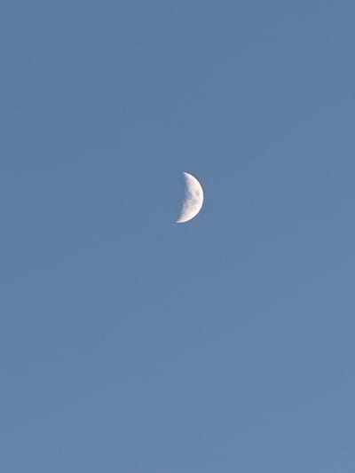 The image shows a crescent moon against a clear blue sky. The moon is partially illuminated, with the visible portion appearing white and cratered. The sky is a uniform light blue, with no clouds or other celestial bodies visible. There are no man-made objects or structures, people, or animals in the image. The scene is tranquil and vast. The provided location of Primorsko-Akhtarsk, Russia, is not discernible from the image itself.