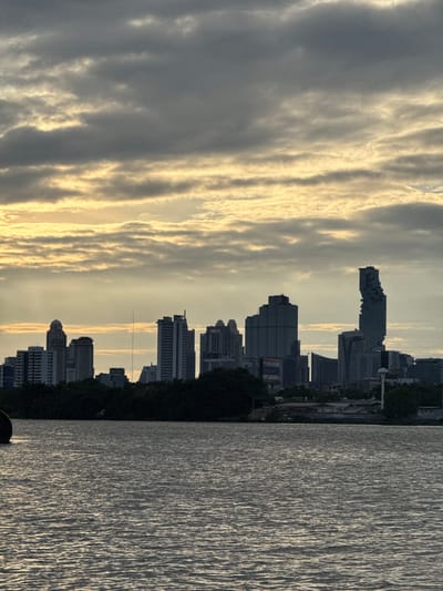 The image depicts a cityscape viewed from across a body of water. The sky is overcast with dramatic clouds, illuminated by the golden hues of either sunrise or sunset. A dense line of trees forms a dark silhouette between the water and the city skyline. The skyline is dominated by numerous tall buildings, including several skyscrapers of varying architectural styles. One particularly distinctive skyscraper on the right has a unique, angular, and somewhat fragmented appearance. The water in the foreground is rippled, reflecting the muted light of the sky. A large, dark, round object, possibly a buoy or part of a floating structure, is partially visible on the left edge of the frame. The scene suggests a metropolitan area with a significant waterfront. The location is identified as Bang Kachao Subdistrict, Thailand.
