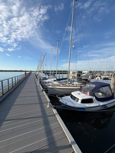 The image shows a pier in the city of Ribnitz-Damgarten, Germany. The pier is made of grey wooden planks and has metal railings on the left side. Several sailboats and a small motorboat are docked along the pier on the right. The boats are of various sizes and colors, with some having blue sails. The water is dark and reflects the boats and the sky. The sky is blue with scattered white clouds, and the sun is shining, casting shadows on the pier. In the distance, there is a body of water and a treeline. One person, wearing a red life jacket, is visible standing on the pier in the middle distance, facing away from the camera. Another person is visible further down the pier. The overall scene depicts a calm day at a marina.