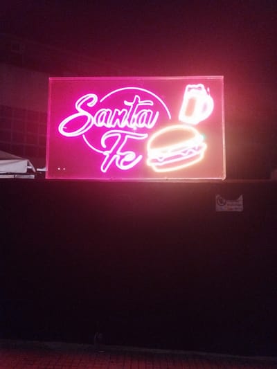 The image shows a neon sign with the words "Santa Fe" in cursive script, along with a burger and a beer mug, likely indicating a restaurant or bar. The sign is illuminated with bright pink and orange light. The background is dark, suggesting it's nighttime or a dimly lit interior. The sign is positioned on top of a dark surface. Based on the prompt, the location is in Urbanització El Carmel, Spain, although there is no geographical information in the image that would allow the identification of the city.