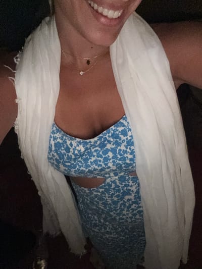 A high-angle, close-up photograph depicts an individual, likely female, from the upper torso to the lower face, situated in Psarou, Greece. The subject is smiling, revealing a row of white teeth. She wears a blue and white floral-patterned dress featuring lateral cut-outs at the waist. A light-colored, presumably white, sheer scarf with frayed edges is draped over her shoulders. Two delicate gold-toned necklaces are visible on the décolletage: one bearing a small, clear, faceted pendant, and the other displaying a small, dark, charm, consistent with a Hamsa symbol. The background is uniformly dark and lacks distinguishing features.