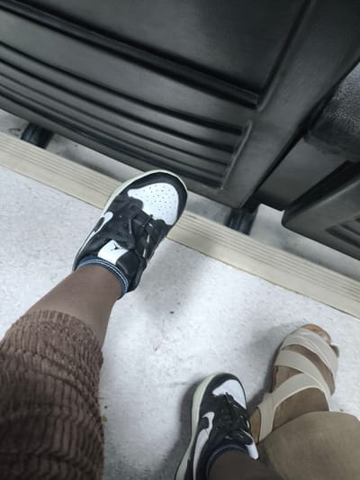 This image captures a low-angle, downward perspective of the lower extremities of two individuals in Kızılbaş, Northern Cyprus / Cyprus. The scene takes place on a light grey, speckled floor surface. A light beige horizontal strip traverses the mid-ground, marking a transition or structural element. In the upper background, a dark grey or black, heavily textured object with prominent horizontal ribs, likely a piece of molded plastic or metal furniture such as the underside of a bench, is visible, supported by two dark, cylindrical feet.
One individual's left foot, positioned on the left, wears a black and white Nike Air Jordan 1 low-top sneaker featuring a white toe box, black leather overlays, black laces, a white Nike Swoosh, and a white Jumpman logo on the tongue. A dark blue sock with a white stripe is visible above the shoe collar. This individual's left lower leg is covered by a brown, deeply ribbed fabric garment. Their right foot, partially visible in the lower center-right, also wears a matching black and white Nike Air Jordan 1 low-top sneaker.
A second individual's right foot is visible on the far right, clad in a light beige or cream-colored open-toed sandal with multiple crisscrossing straps, one of which wraps around the big toe. This individual's lower leg is covered by a light tan fabric. Both individuals appear to be seated or standing in close proximity.